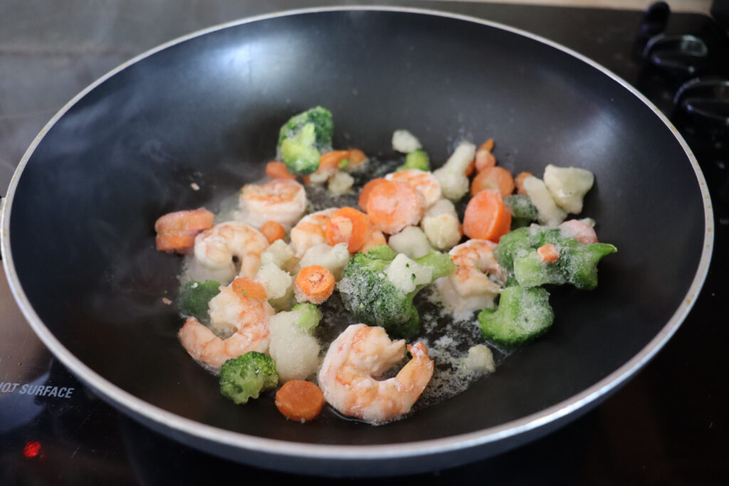 Pink shrimp and frozen vegetables cooking in a deep pan