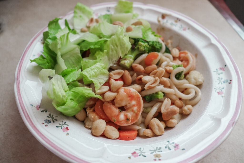 A bowl of stir fried udon noodles with shrimp, veggies, and fresh lettuce.